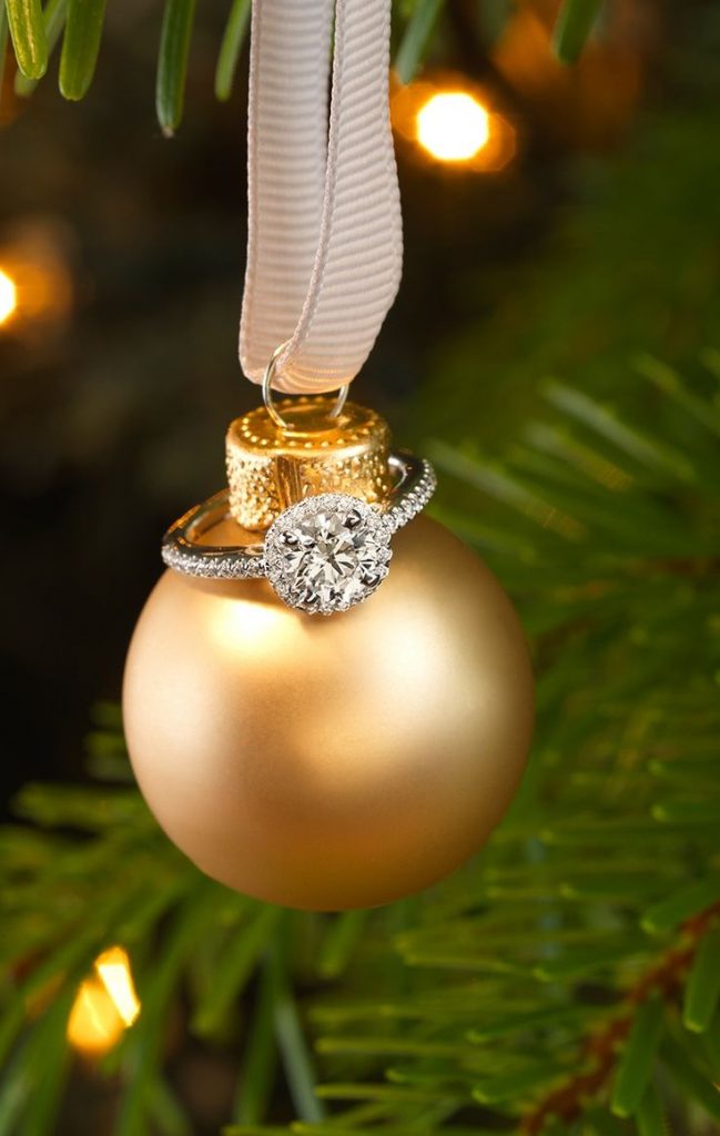 Anillo de pedida con diamantes colgando en una bola de navidad. Anillo de compromiso de oro blanco con adorno de navidad de un árbol con luces navideñas